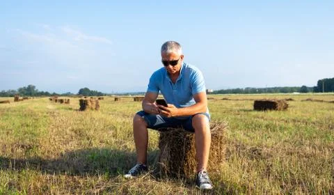 A man sitting in a field with a phone on a haystack. Stock Photos