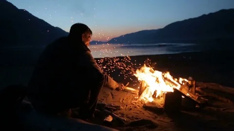 A man is sitting by a fire and throwing logs into it late at night Stock Footage 73349415