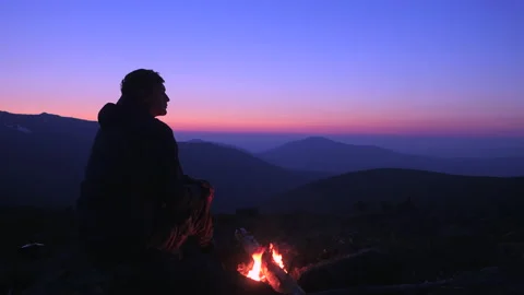 A man is sitting by a fire in the mountains looking at the night sky and warm Stock Footage 135897947
