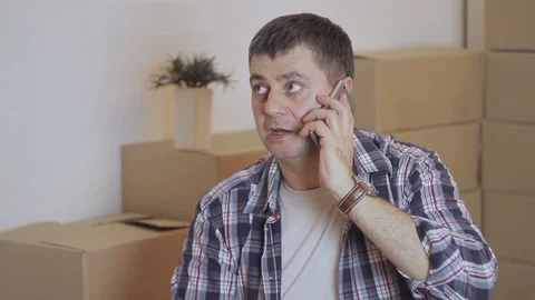 A man is sitting on the floor and talking on the phone in a room with cardboard Stock Footage 86669960