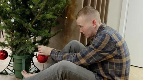 Man sitting on floor, decorating small christmas tree with red baubles and Fotos Stock