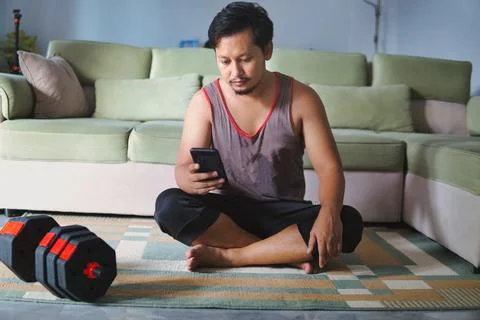 Man Sitting on Floor at Home Using Phone Beside Dumbbells Stock Photos
