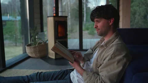 Man sitting on the floor by the sofa and casually reading a book in front of the Vídeos de archivo 245991650