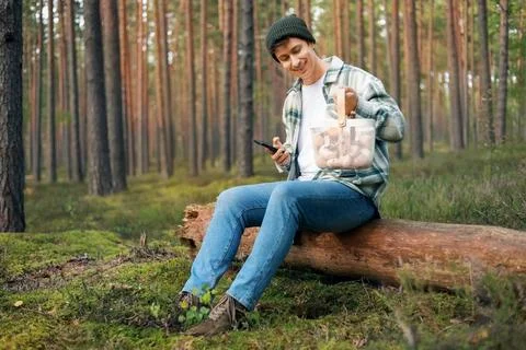 Man sitting on a forest log holding the smartphone and a basket full of wil.. Stock Photos