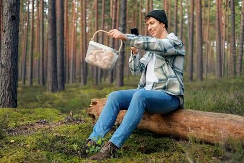 Man sitting on a forest log takes a smartphone photo of his autumn harvest... Foto stock