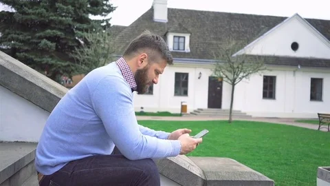 Man sitting in front of big house, browsing smartphone, look at camera steadycam Vídeos de archivo 74345013