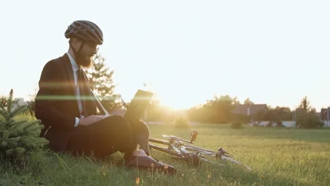 Man sitting on a grass at the park working on a laptop, wearing a helmet Video stock 163353769
