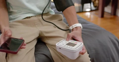 Man sitting on gray bed, pressing BP monitor, holding cuff, checking phone for Stock Footage 339679466