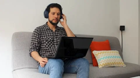 Man sitting on a grey couch listening music on a laptop over his legs Vídeo Stock 340583364