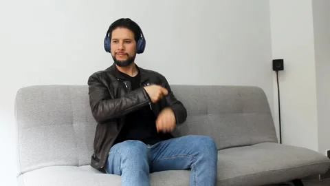 Man sitting on a grey couch listening music on a headset and doing drums air Vídeo Stock 340583436