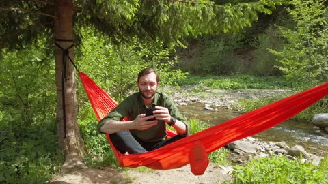 Man sitting in a hammock atnature using smartphone swiping screen up. Relaxing Stock Footage 131899609