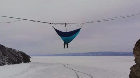 Man sitting in a hammock mounted at high altitude over a frozen lake. Stock Footage 107943228