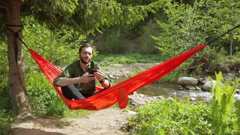 Man sitting in a hammock at the nature talking on smartphone. Scene of relaxing  Stockbeeldmateriaal 131915396