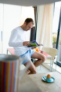 Man sitting on kitchen table, reading magazine, cup and boiled egg in foreground Stock Photos