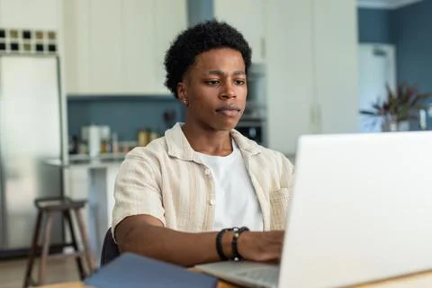 Man sitting at kitchen table using silver laptop, wearing beige shirt and black 写真素材
