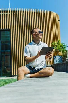 Man sitting on a ledge using a tablet in a sunny outdoor space with a woode.. Stock Photos