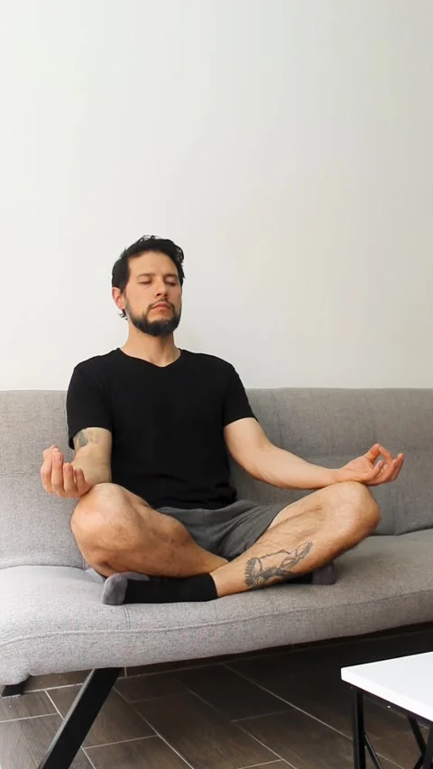 A man sitting at the left side of a couch in the lotus pose meditating Video stock 340583550