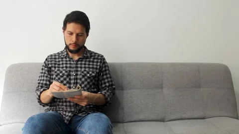 Man sitting at the left side of a grey couch solving crosswords puzzles Vídeo Stock 340583448