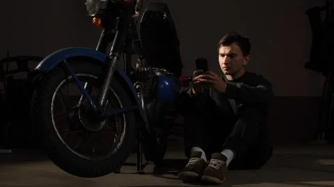 Man sitting next to an old 1970s motorcycle, looking at his smartphone for Stock Footage 342761935