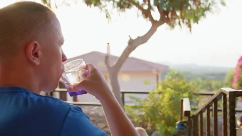 Man is sitting on an open balcony, drinking a refreshing drink from a glass cup Stock Footage 306126414