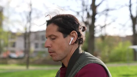 Man sitting in a park spring time listening to music airpods smartphone Stock Footage 237277532