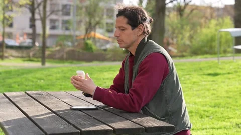 Man sitting in a park spring time listening to music airpods smartphone Stock Footage 237277535