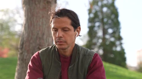 Man sitting in a park spring time listening to music airpods smartphone Stock Footage 237277664