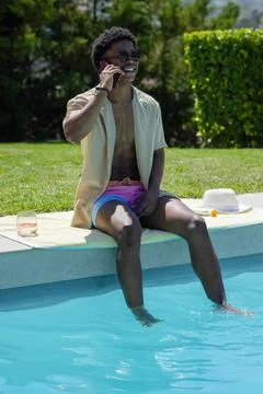 Man sitting on pool edge, talking on smartphone, wearing sunglasses, towel, hat Stock Photos