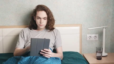 A man is sitting reclining in bed and typing something on a computer tablet Stockbeeldmateriaal 196814405