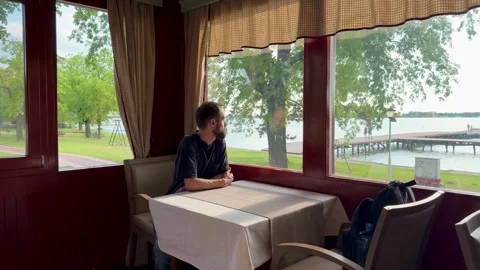 Man Sitting at Restaurant Table by Window Overlooking Lake Palic Vídeos de archivo 336178194