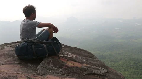 A man sitting on the rock, drinking water and looking to a valley at the bottom Stock Footage 49456366