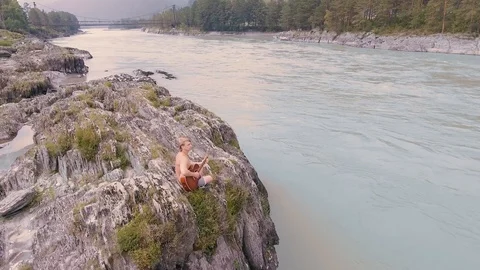 A man sitting on the rocks by a mountain river and playing the guitar. aerial Stock Footage 93733251