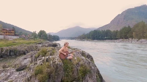 A man sitting on the rocks by a mountain river and playing acoustic guitar Stock Footage 93733482