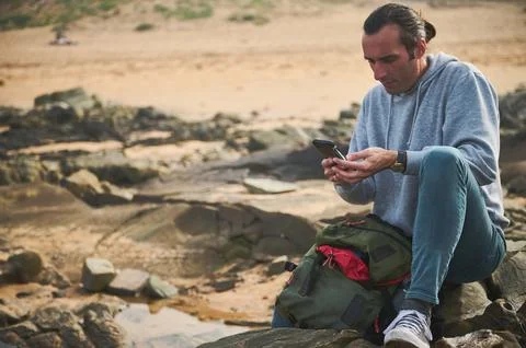 Man sitting on rocky beach using smartphone with backpack Stock Photos