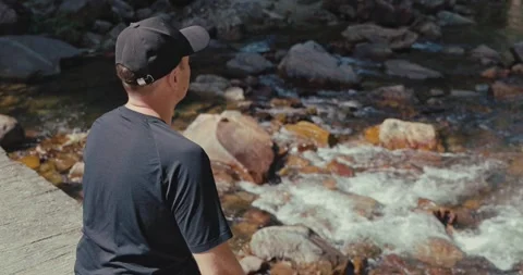 Man sitting by rocky mountain stream in forest 스톡 동영상 334221717
