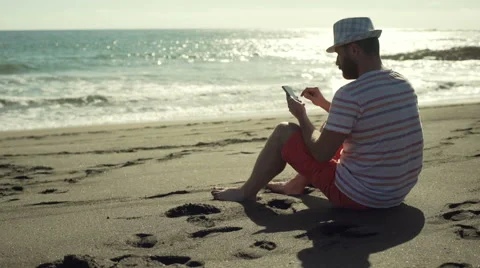 Man sitting on the sandy beach and using smartphone, steadycam shot Vídeos de archivo 60133522