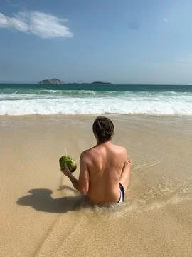 Man sitting on the sandy beach facing the ocean, with a green coconut drink. Stock Photos