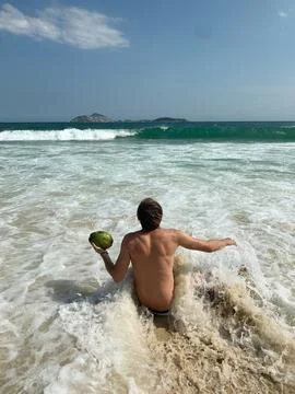 Man sitting on the sandy beach facing the ocean, with a green coconut drink. Stock Photos