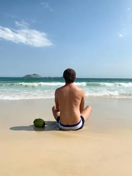 Man sitting on the sandy beach facing the ocean, with a green coconut drink. Stock Photos
