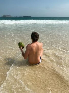 Man sitting on the sandy beach facing the ocean, with a green coconut drink. Stock Photos