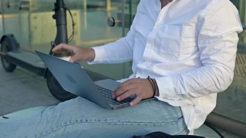 Man is sitting on the sidewalk with a laptop, using a smartphone and drinking Stock Footage 198566702