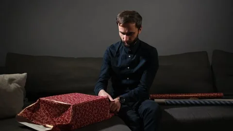 Man sitting on a sofa and wrapping a gift box in bright red paper at home. Stock Footage 340064806
