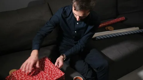 Man sitting on a sofa and wrapping a gift box in bright red paper at home. Stock Footage 340064892