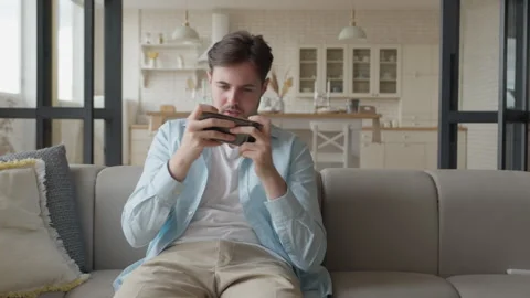 Man Sitting On Sofa Focused On Playing Mobile Game, Immersed In Virtual Battle 動画素材 302450414