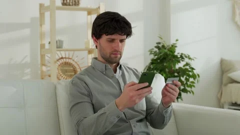 Man sitting on the sofa holding debit card and using smartphone for shopping at Vídeo Stock 150027003