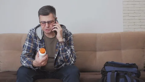 A man sitting on a sofa holds a jar of pills in his hands and uses a mobile Stock Footage 104589487
