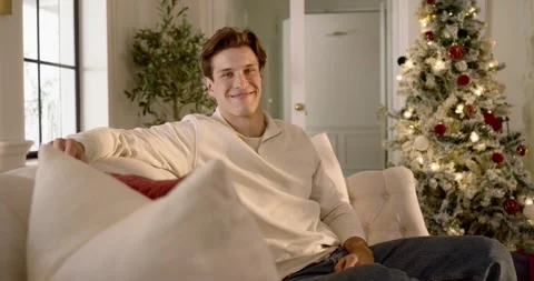 Man sitting on sofa at home beside lit Xmas tree gifts while camera approaching, Stock Footage 344655189