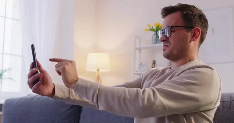 Man sitting on sofa at home in eyeglasses trying to read message on smartphone Stock Footage 311936060