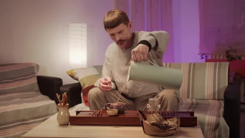 A man is sitting on a sofa at home, pouring boiling water from a thermos into a Stock Footage 145527872