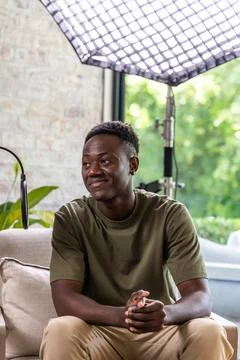Man sitting on sofa in home studio wearing olive T-shirt, beige pants, being lit Stock Photos
